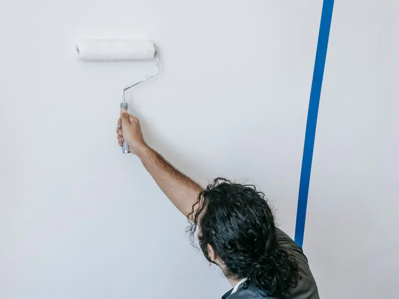 Painter rolling white paint onto an interior wall edged with blue tape